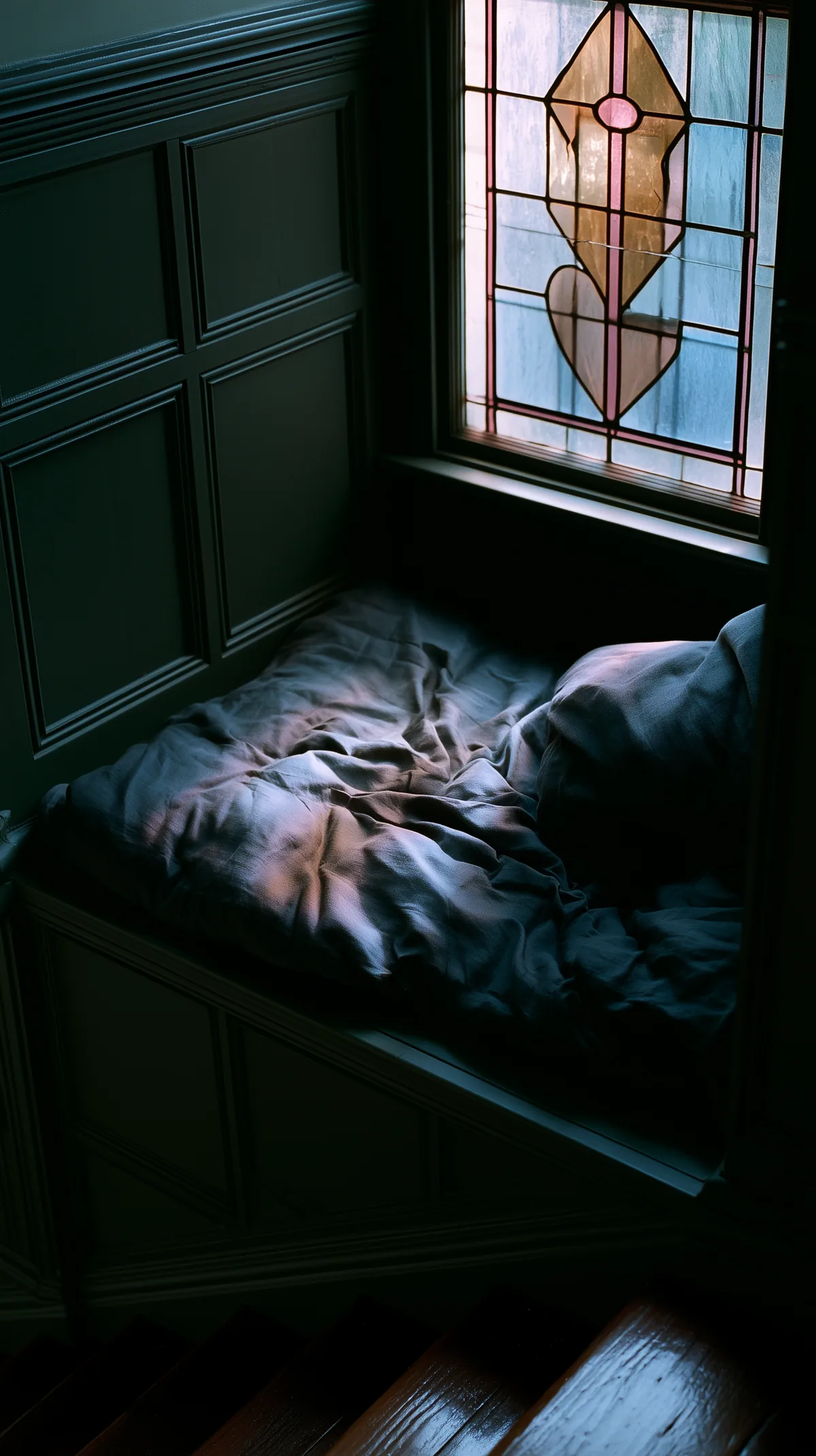 A dark panelled window seat on a staircase landing, rumpled grey linen cushions below a leaded stained glass window in muted rose and amber